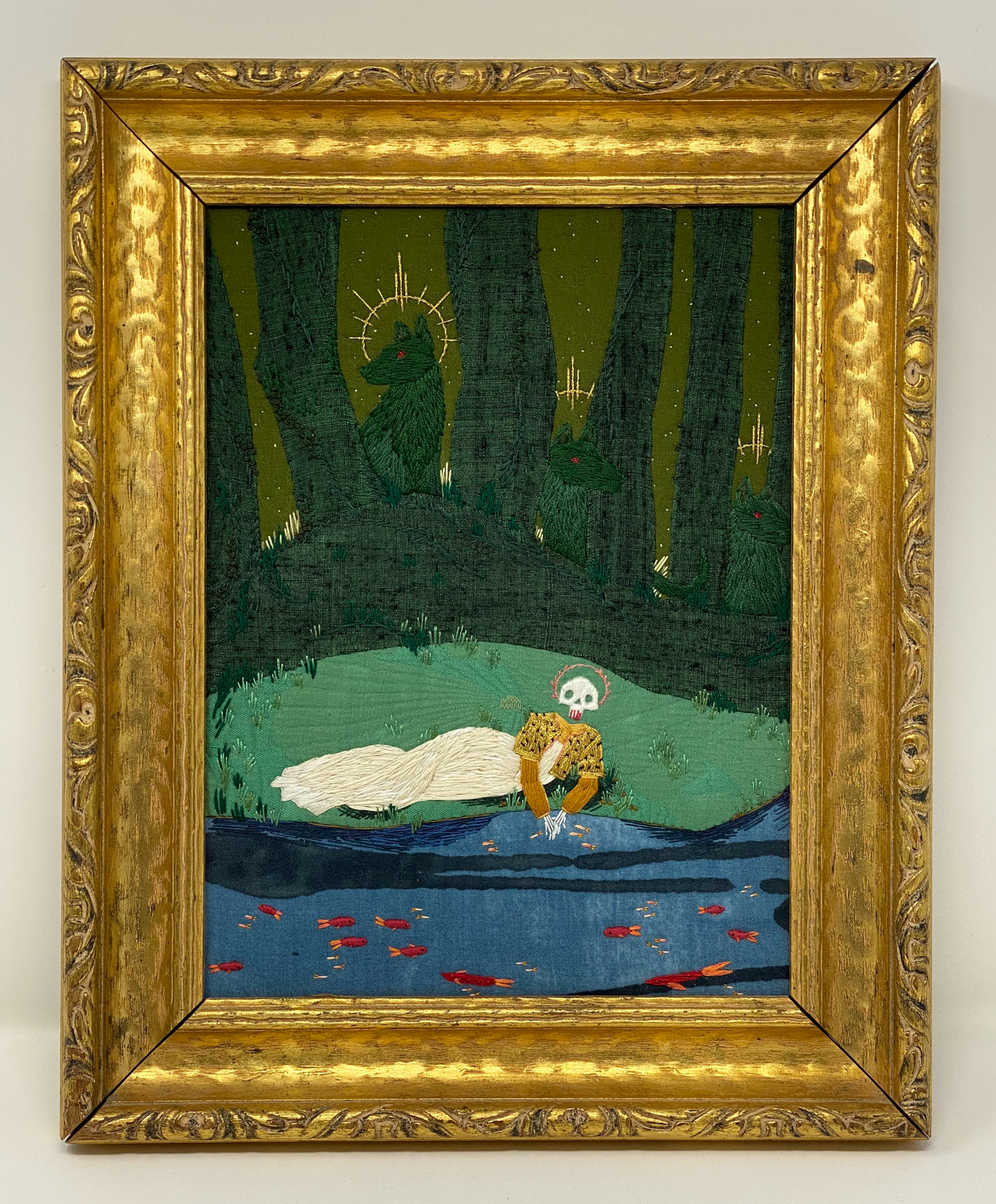 LARGE GOLD FRAME WITH GREEN FABRIC TREES AND HILL. GREEN DOGS ARE HIDDEN IN THE TREES WITH GOLD HALOS AROUND THEM. THERE IS A SKULL FACED PERSON IN A GOLD COAT AND WHITE DRESS LAYING NEXT TO A STREAM WITH RED FISH. THIS IS ALL HAND EMBROIDERY ON FABRIC. 
